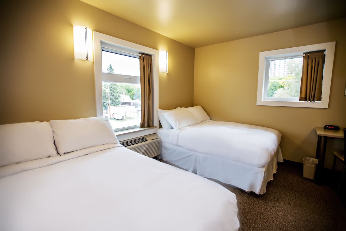 A hotel room with two beds, a window with curtains, a wall-mounted heater, and a small table with a clock.