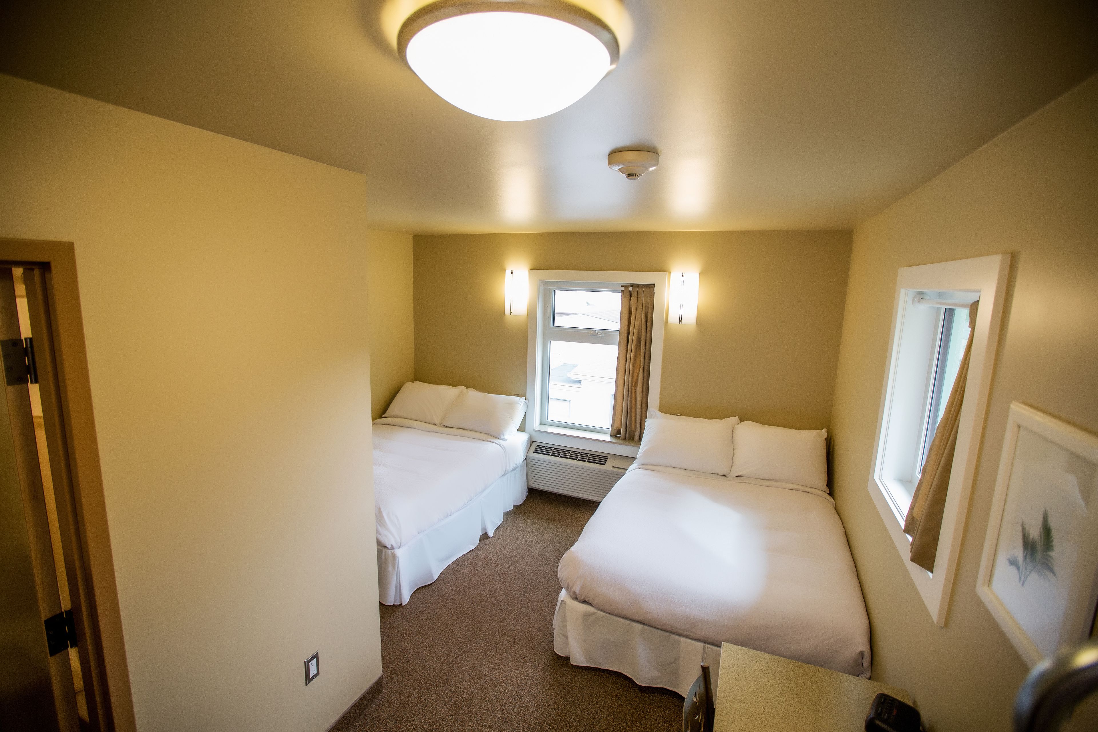 A hotel room with two beds, a window with curtains, a ceiling light, and a framed picture on the wall.