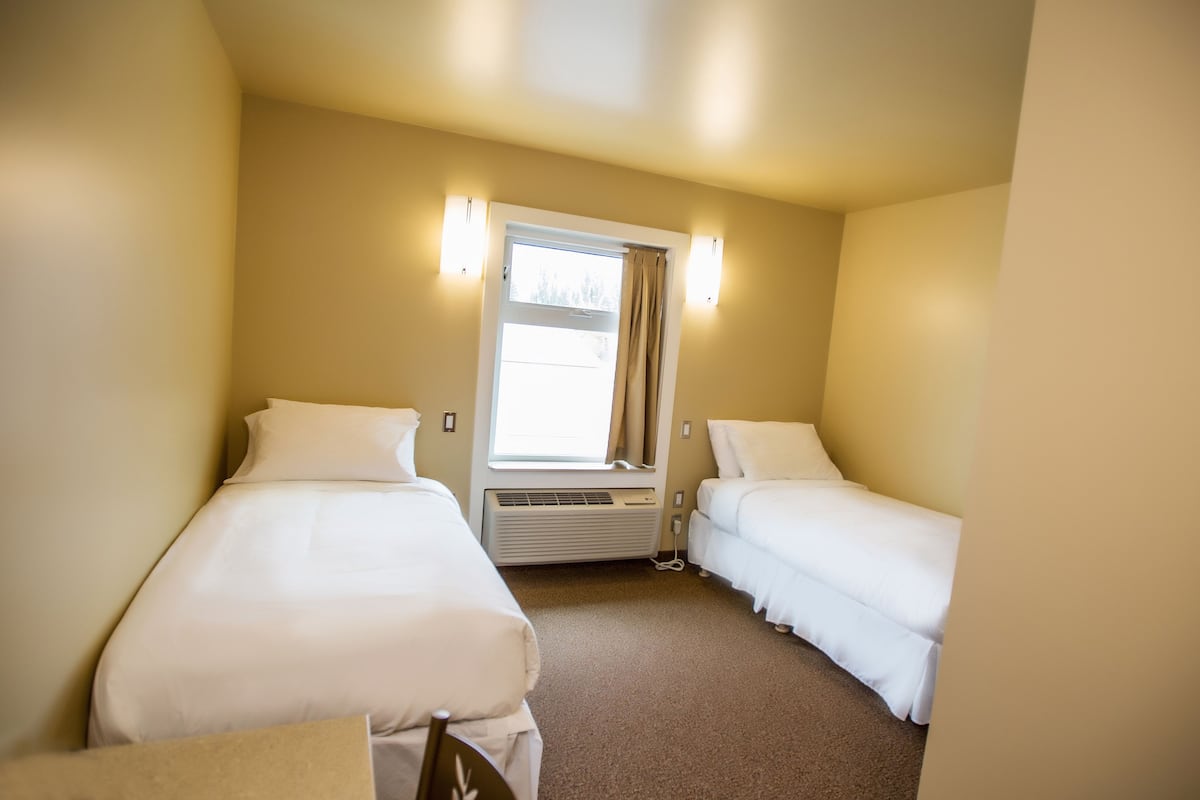 A hotel room with two single beds, a window with curtains, wall-mounted lights, and a heater.