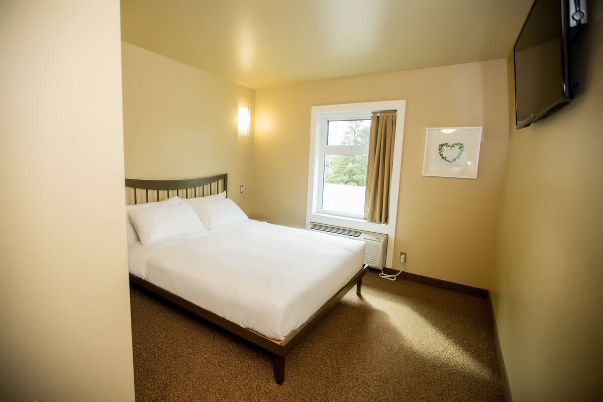 A neatly made bed in a room with a window, a wall-mounted TV, and a small picture frame.