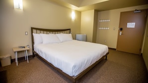 Habitación de hotel con una cama individual, una mesita de noche pequeña, un cesto de basura, una lámpara fijada a la pared y una puerta con perchero.