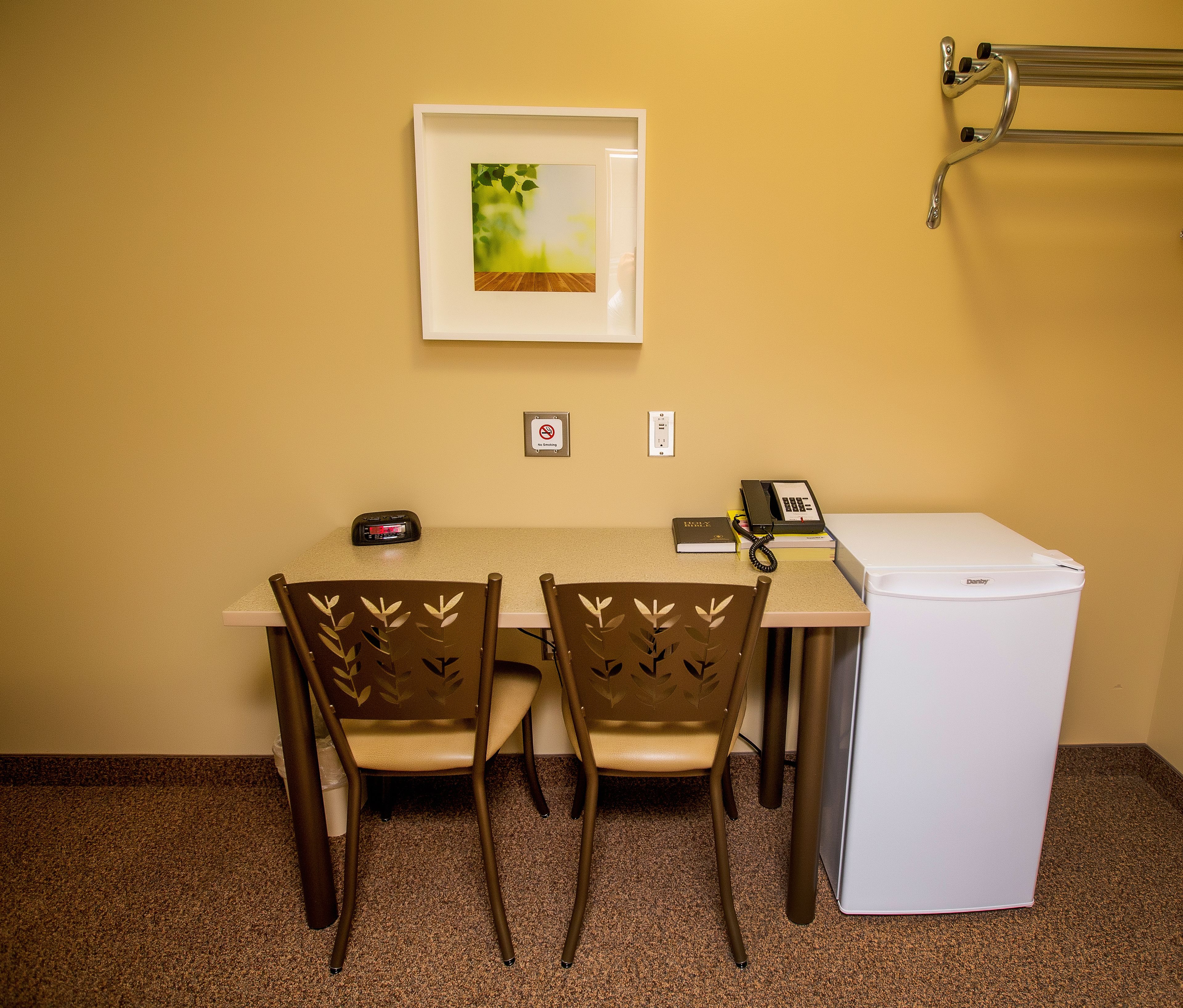 A small office with a desk, two chairs, a refrigerator, a phone, a clock, and a framed picture on the wall.