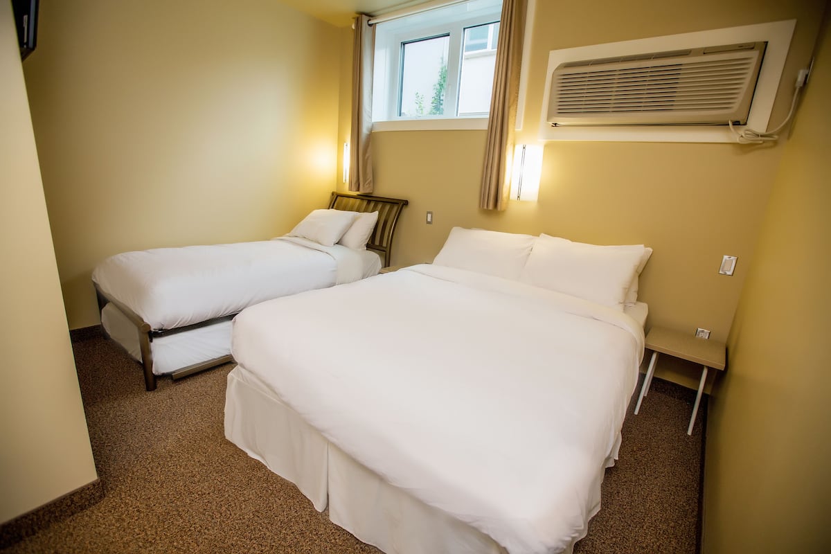 A hotel room with two beds, a window with curtains, an air conditioning unit, and a small table.