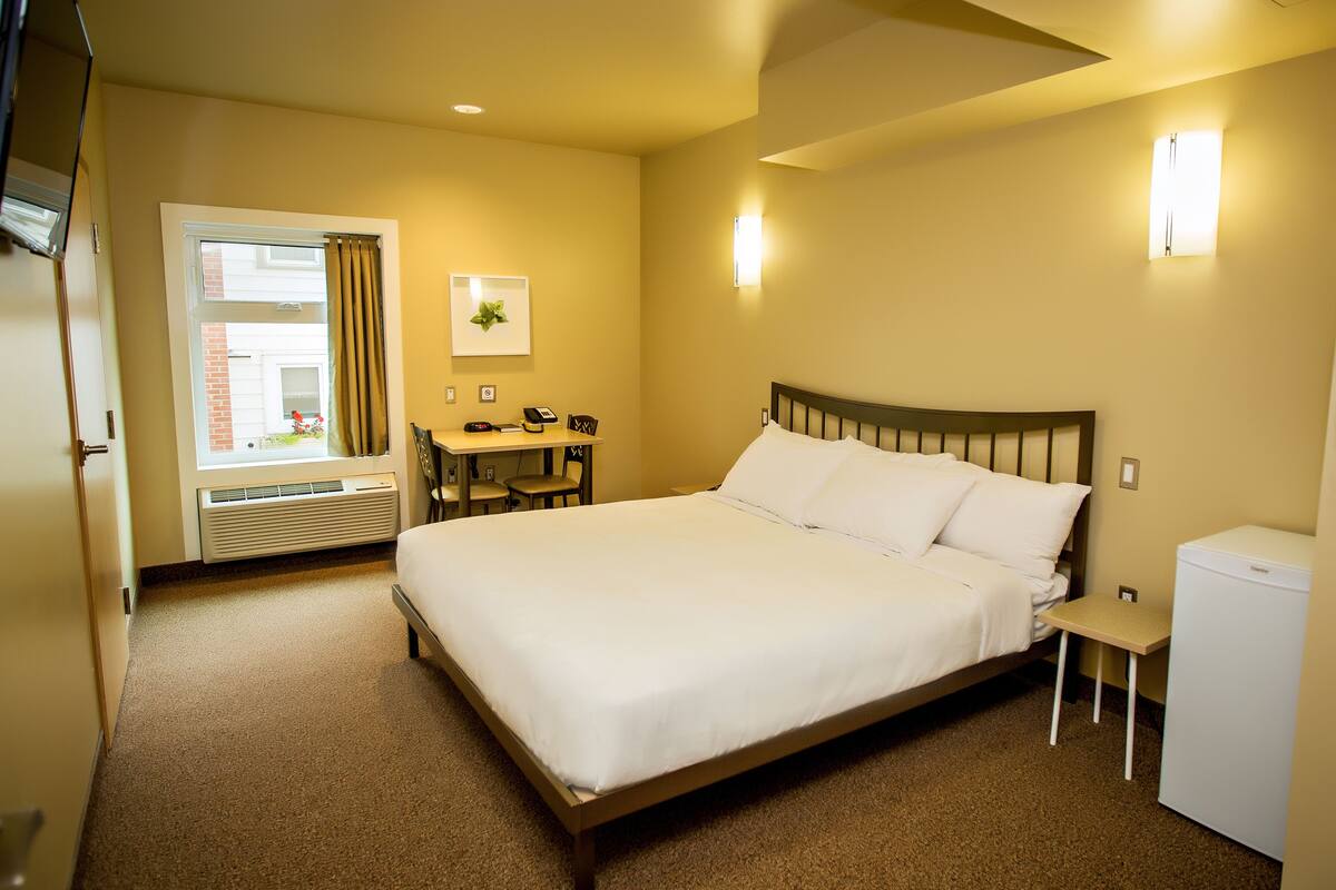 A hotel room with a large bed, a desk with a chair, a small table, and a refrigerator.