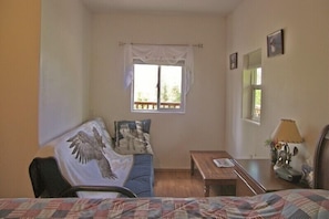 A bedroom with a bed, a window with curtains, a wooden table, and a lamp. - Grace and Bill's Freedom Hills (Talkeetna)