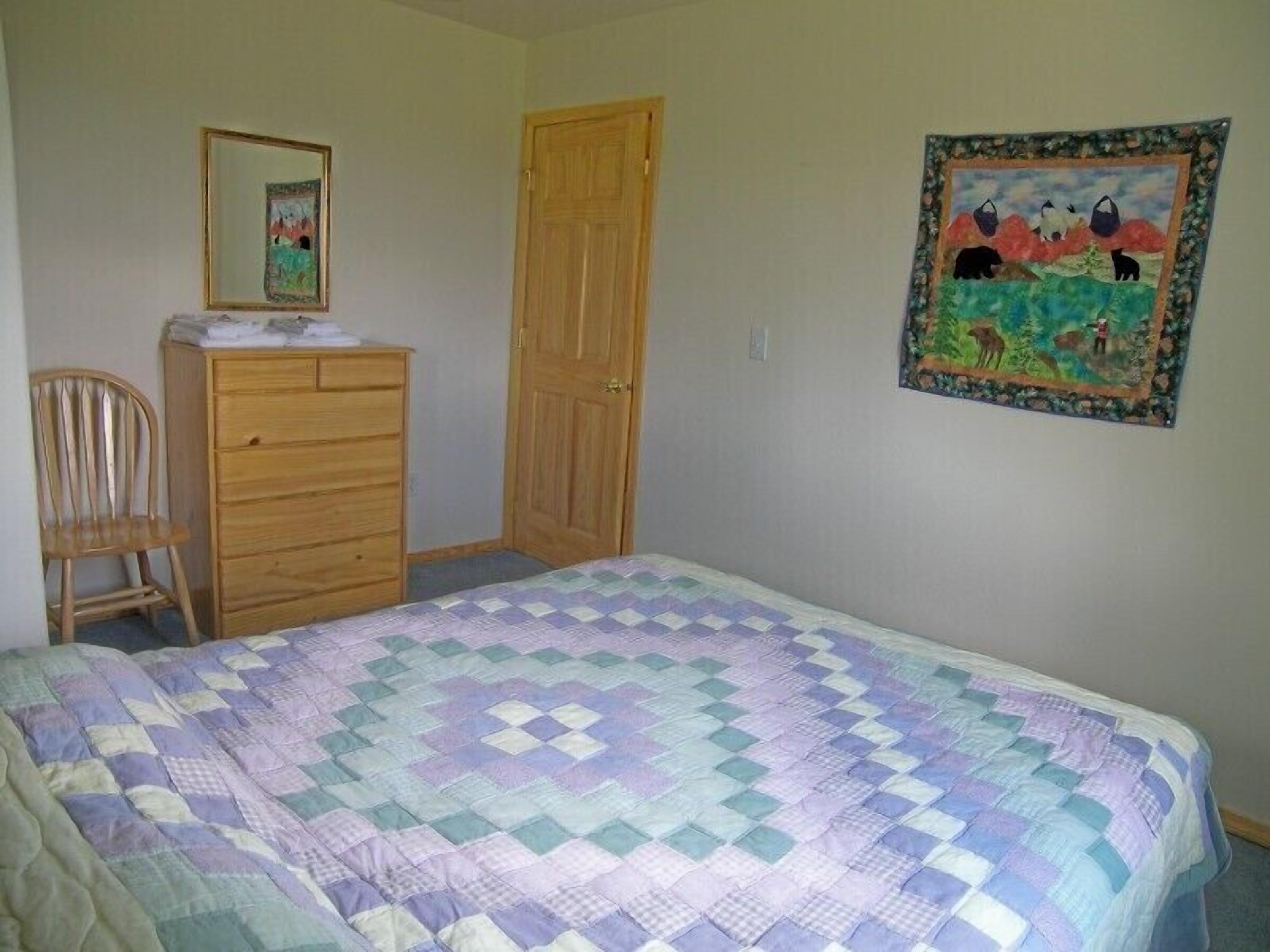 A bedroom with a bed, a wooden dresser, a chair, a door, and a wall decoration.