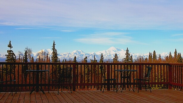 A wooden deck with metal chairs and tables, overlooking a snowy mountain range and autumn-colored trees. - Grace and Bill's Freedom Hills (Talkeetna)