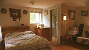 A bedroom with a large bed, wooden dresser, and a chair. There is a window with curtains, a door leading to a bathroom, and a mirror on the wall. - Grace and Bill's Freedom Hills (Talkeetna)