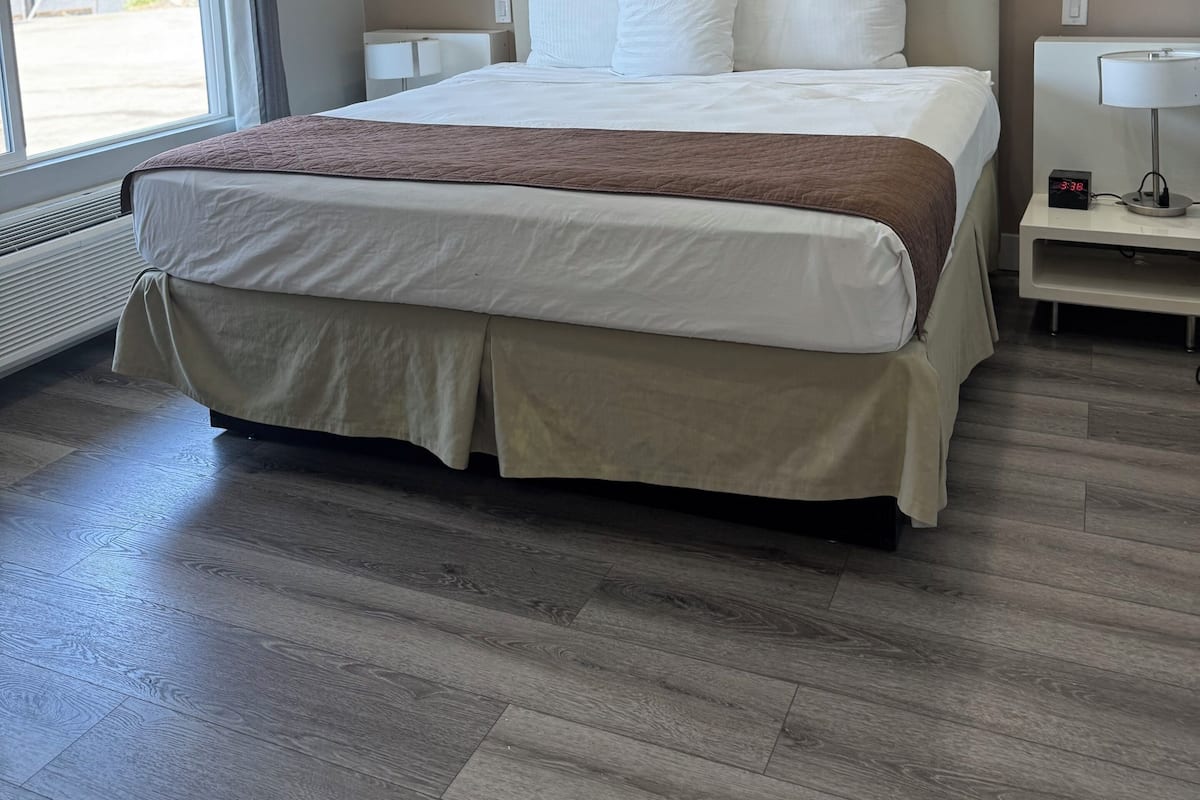 A modern hotel room with a large bed, a bedside table with a lamp, and a view of the outside through a large window.