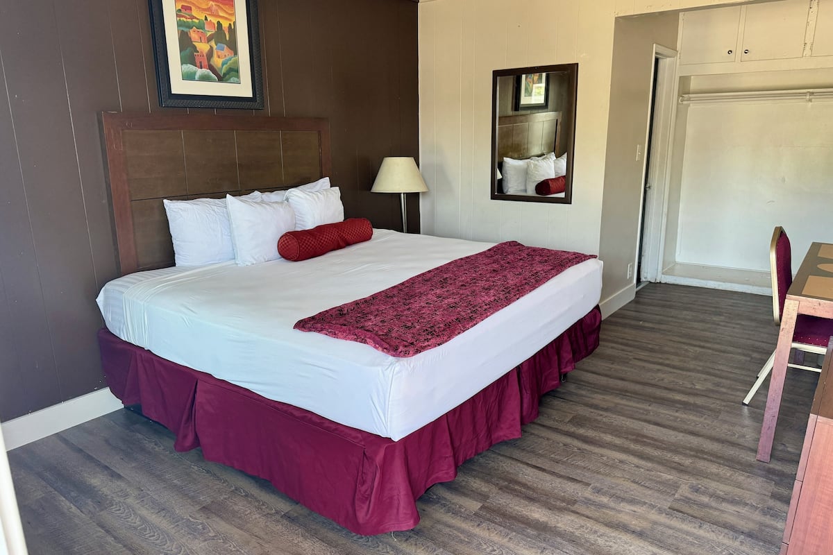 A hotel room with a large bed, a wooden headboard, a red and white bedspread, a red throw blanket, a nightstand with a lamp, a framed picture on the wall, and a wooden desk with a chair.