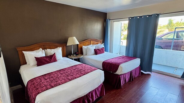 Habitación de hotel con dos camas, una mesita de noche con lámpara, ventana con cortinas y vista a un estacionamiento.