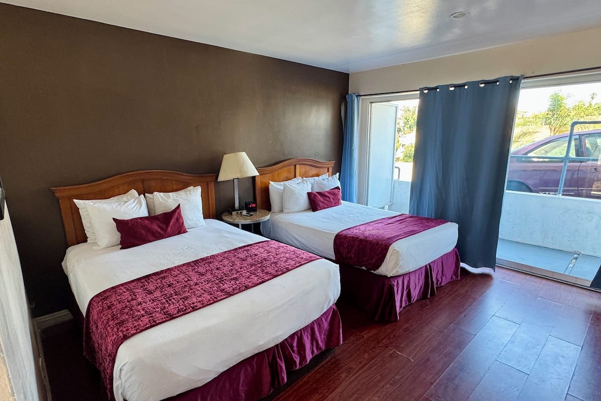 A hotel room with two beds, a nightstand with a lamp, a window with curtains, and a view of a parking area.