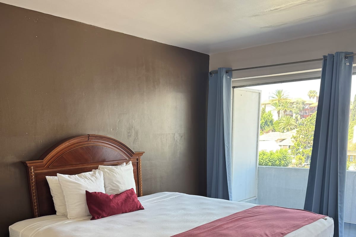 A large bed with a wooden headboard, white and red bedding, and a view of greenery outside through a window with curtains.