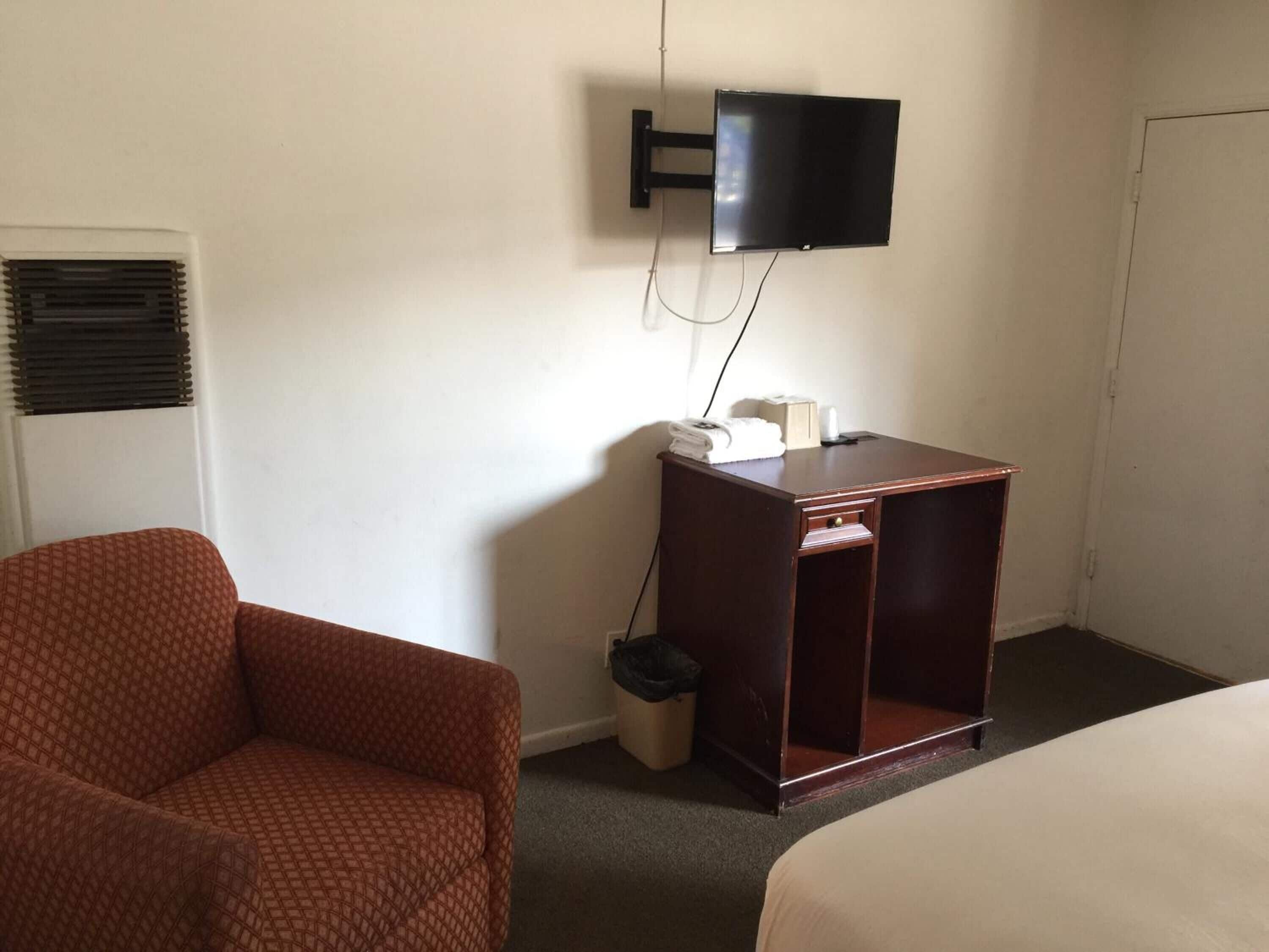 Une chambre d’hôtel avec un lit simple, une table de chevet en bois, un téléviseur fixé au mur, un fauteuil à motifs et une fenêtre avec des stores.