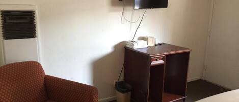 A hotel room with a single bed, a wooden nightstand, a television mounted on the wall, a patterned armchair, and a window with blinds.