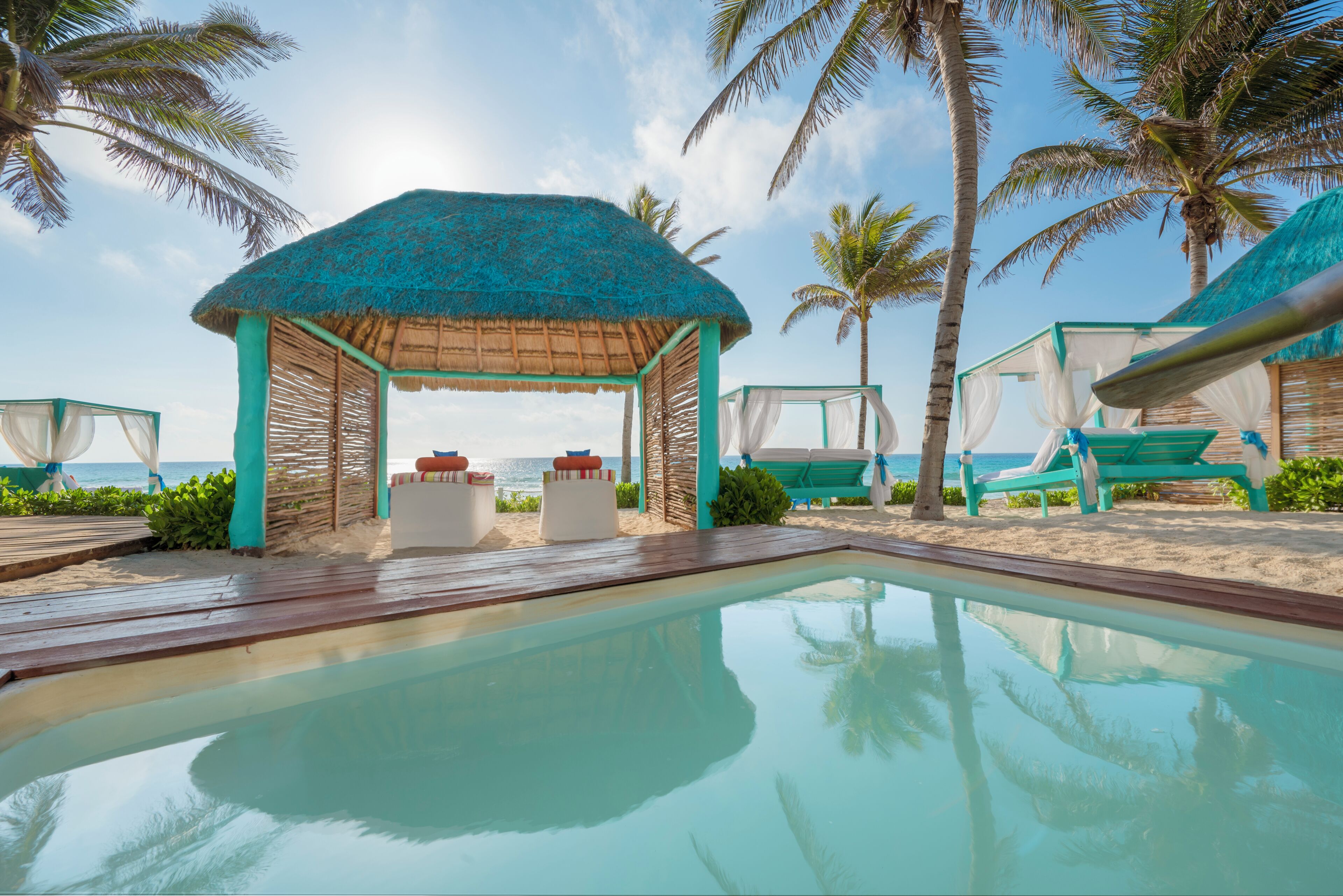 A pool area with a thatched-roof gazebo, lounge chairs, and palm trees.
