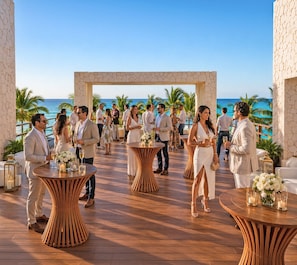 An outdoor event with round wooden tables, guests, and a view of the ocean. - The Pyramid Cancun by Oasis - Gourmet Premium All Inclusive (Cancun)
