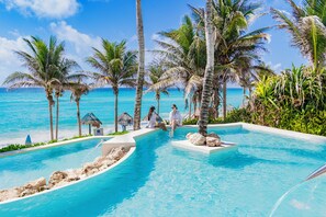 A pool area with clear blue water, surrounded by palm trees and a view of the ocean. - The Pyramid Cancun by Oasis - Gourmet Premium All Inclusive (Cancun)