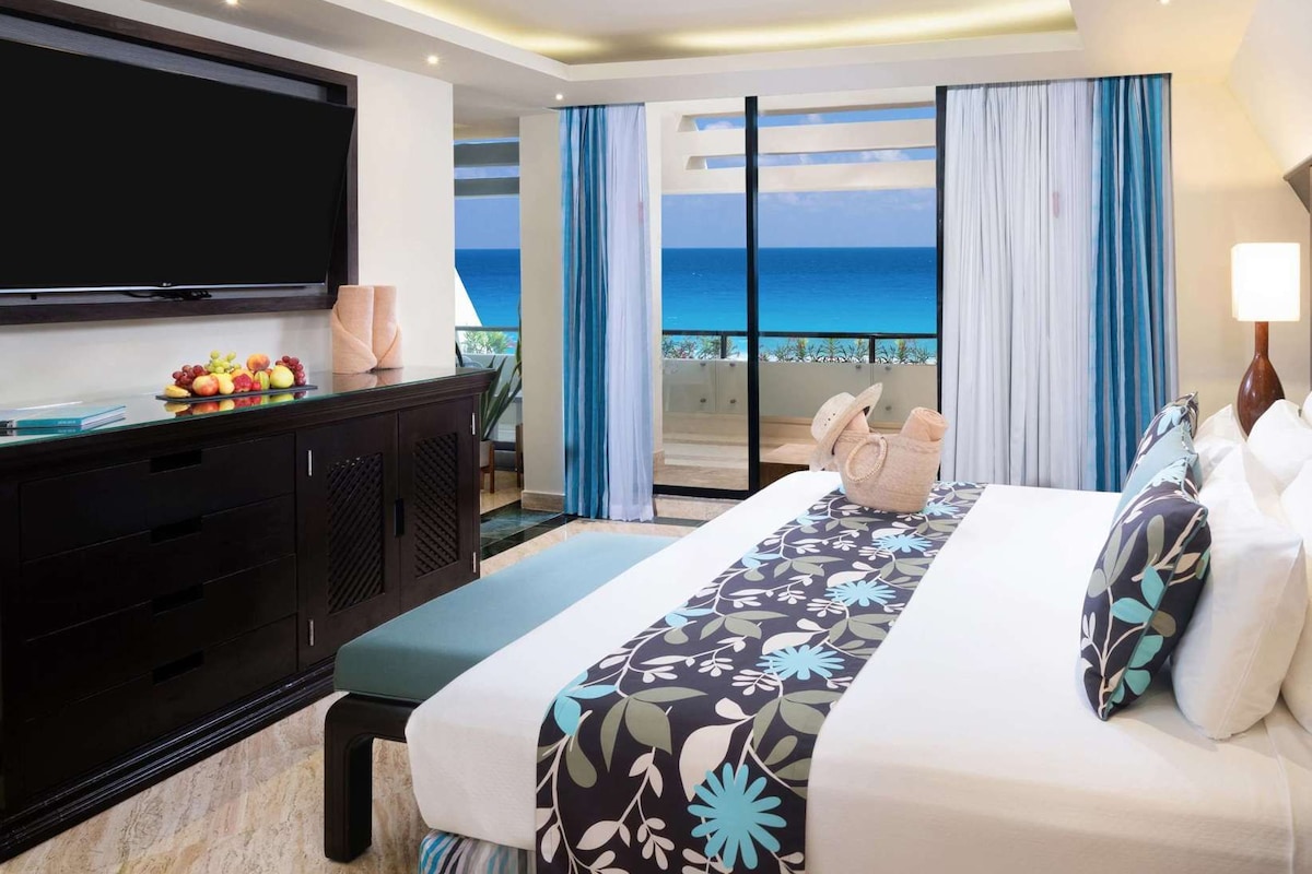 Une chambre d’hôtel avec un lit, une télévision, un bureau avec des fruits et une vue sur l’océan.