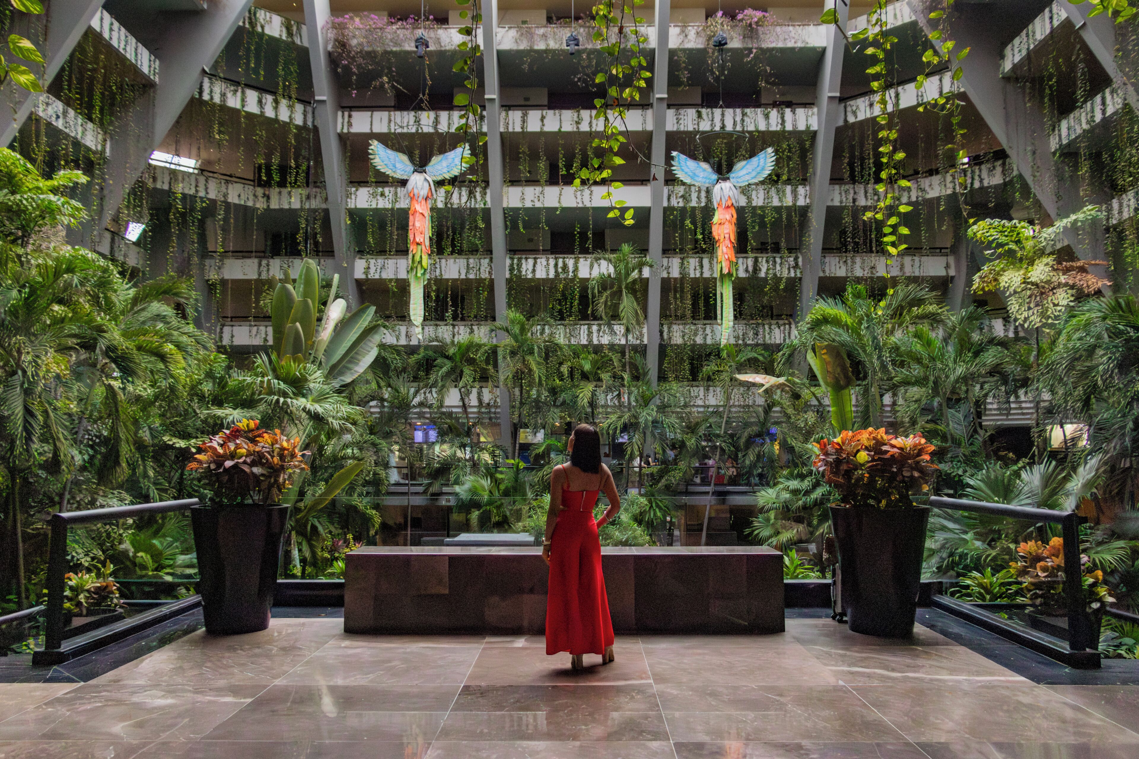 A spacious atrium with a central walkway, surrounded by lush indoor plants and hanging decorations.