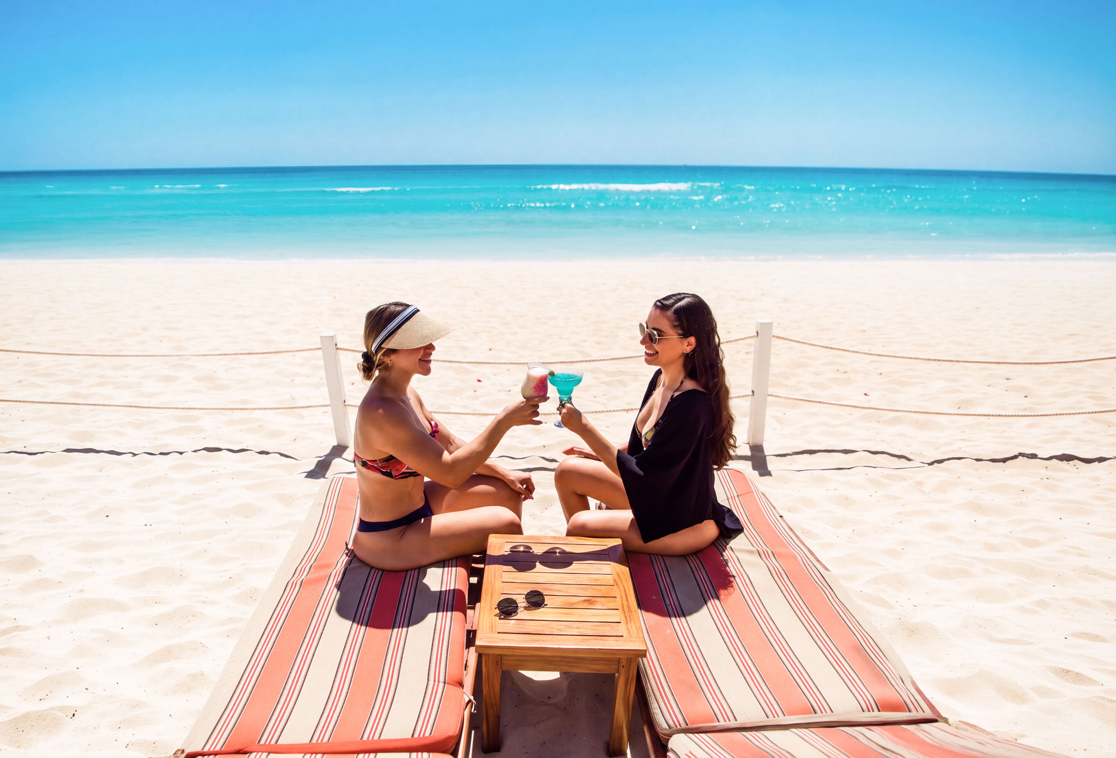 Two people sitting on a striped beach mat with a wooden table between them, facing the ocean.