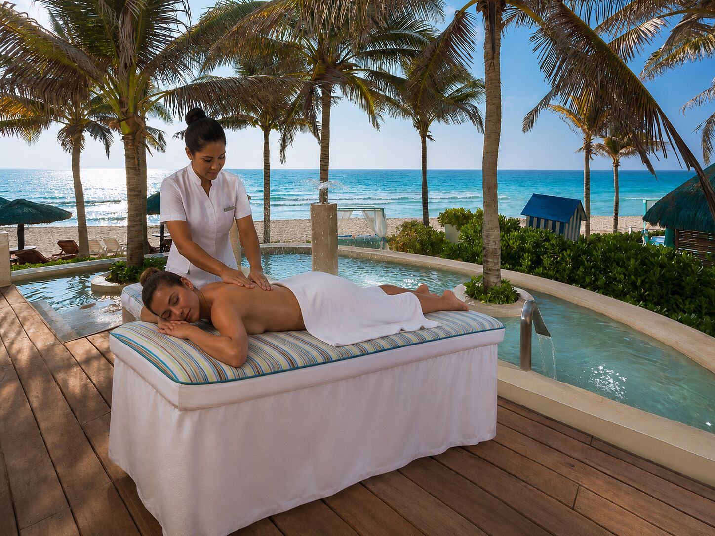 Zona de spa con una mesa de masaje, una persona recibiendo un masaje y una piscina con vista a la playa y el océano.