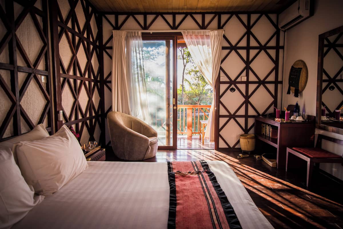 A bedroom with a bed, a chair, a wooden floor, a bookshelf, and a view through a glass door.