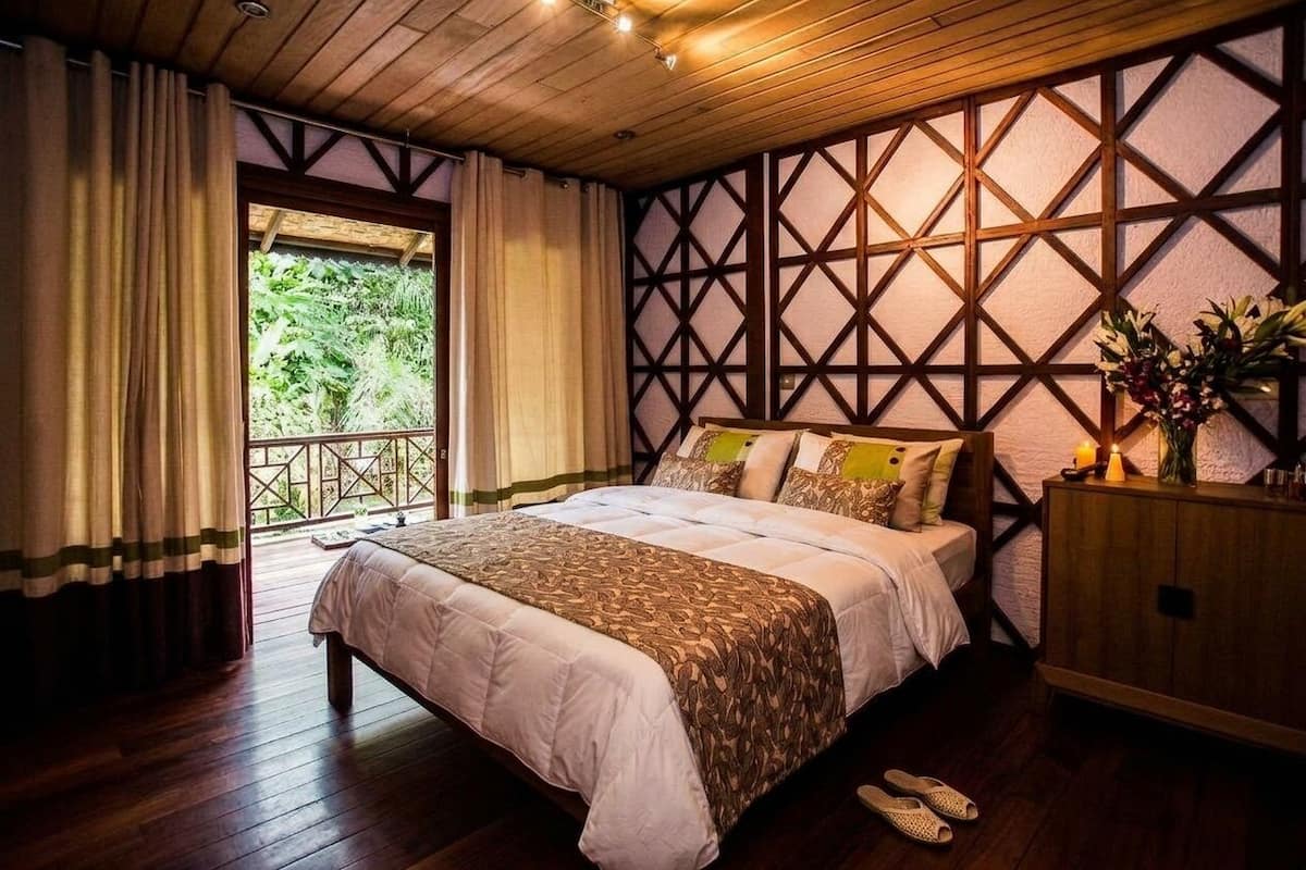 A bedroom with a wooden ceiling, a large bed with patterned bedding, a wooden cabinet, and a view of greenery outside.