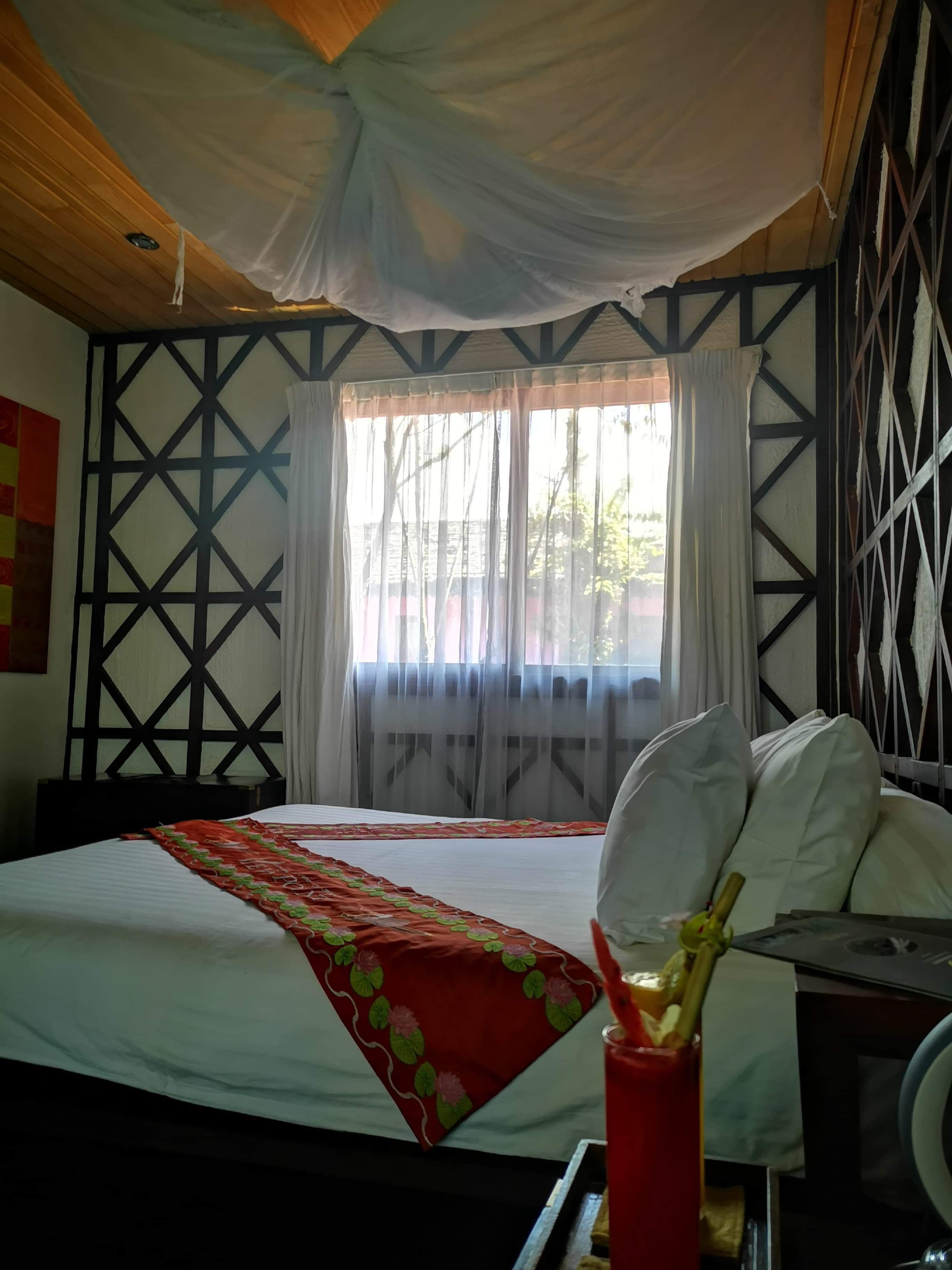 A hotel room with a bed, a nightstand, a red vase with a plant, and a window with curtains.