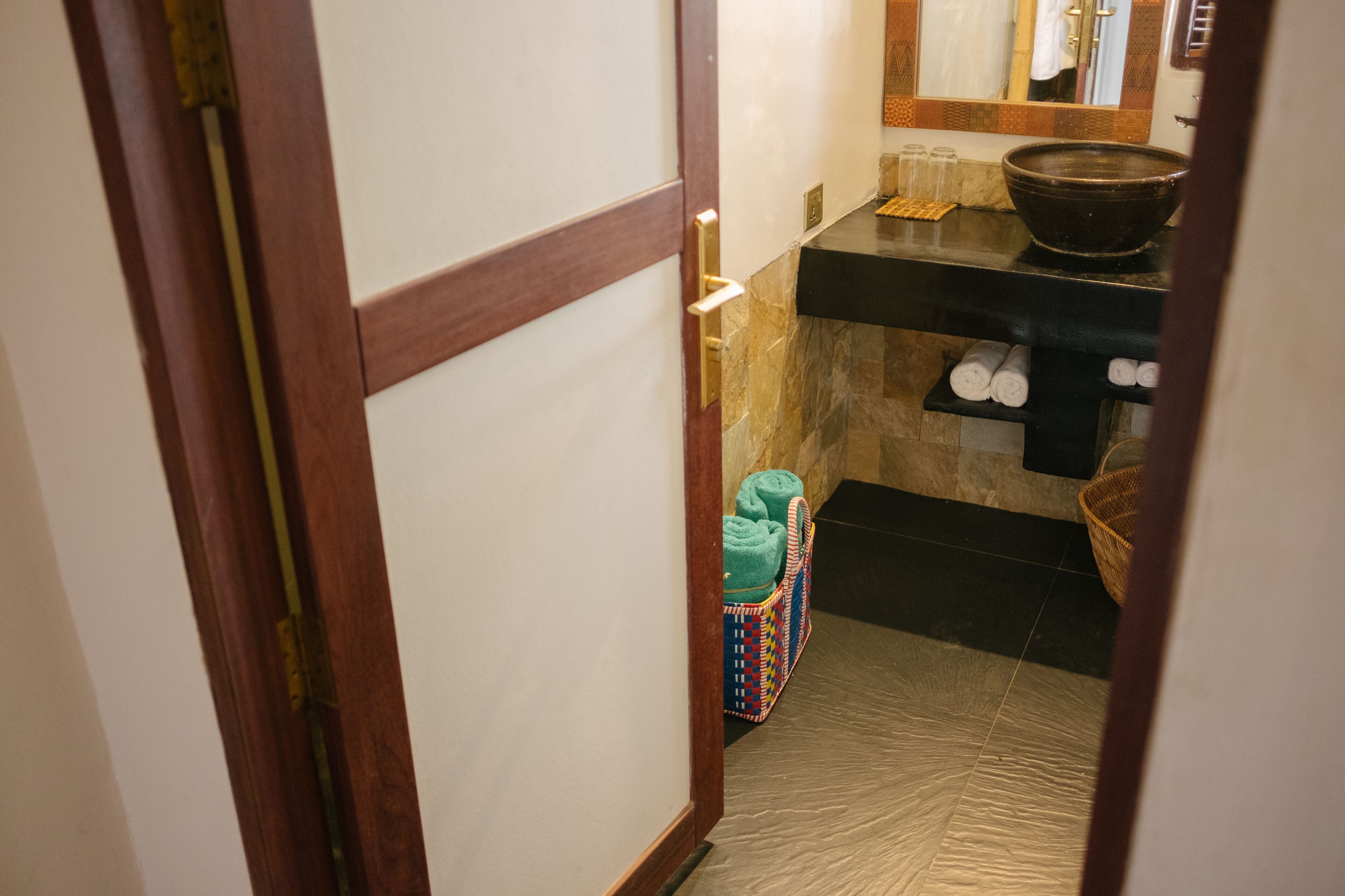 A narrow hallway with a wooden door, a tiled floor, a black countertop with a bowl, and a basket with towels.
