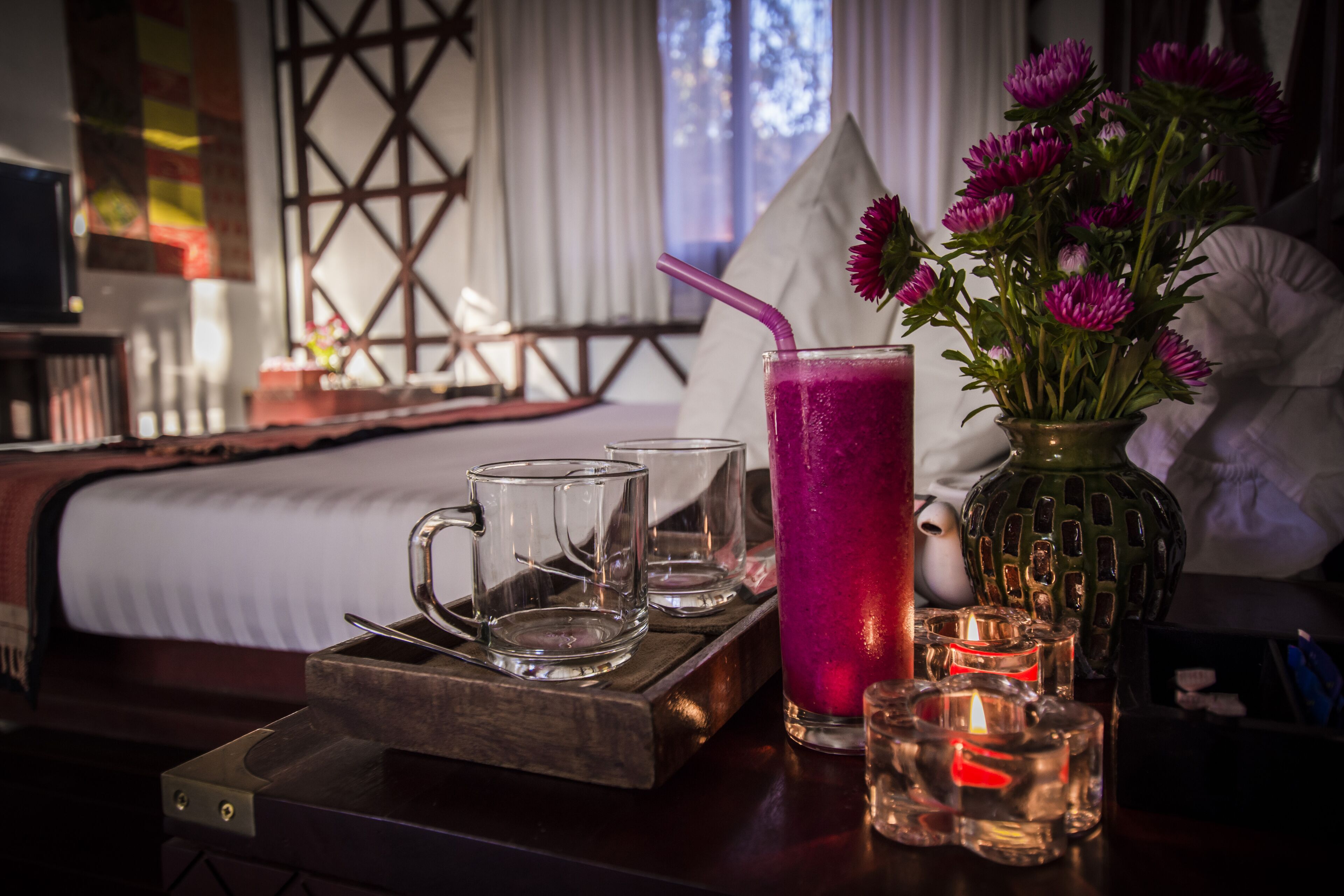Una habitación con una cama, una mesita de noche con una bandeja que contiene vasos y una bebida, un florero con flores y velas.
