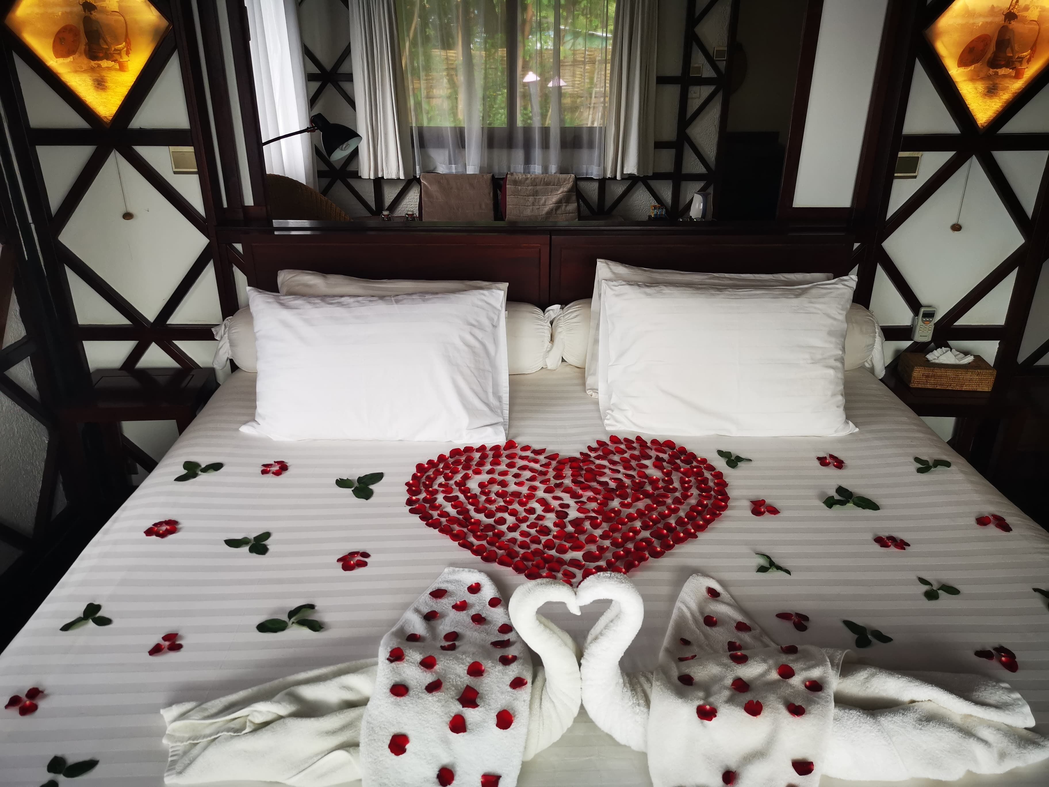 Een opgemaakt bed met een hartvormige decoratie van rode rozenblaadjes en twee zwaan-handdoeken.