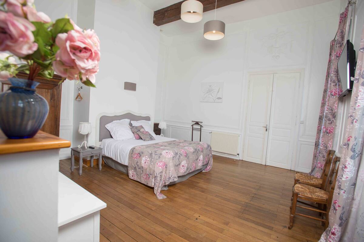 Une chambre avec un plancher en bois, un lit recouvert d’une couvre-lit à motifs floraux, une table de chevet, une lampe, un vase avec des fleurs, une chaise et un téléviseur fixé au mur.