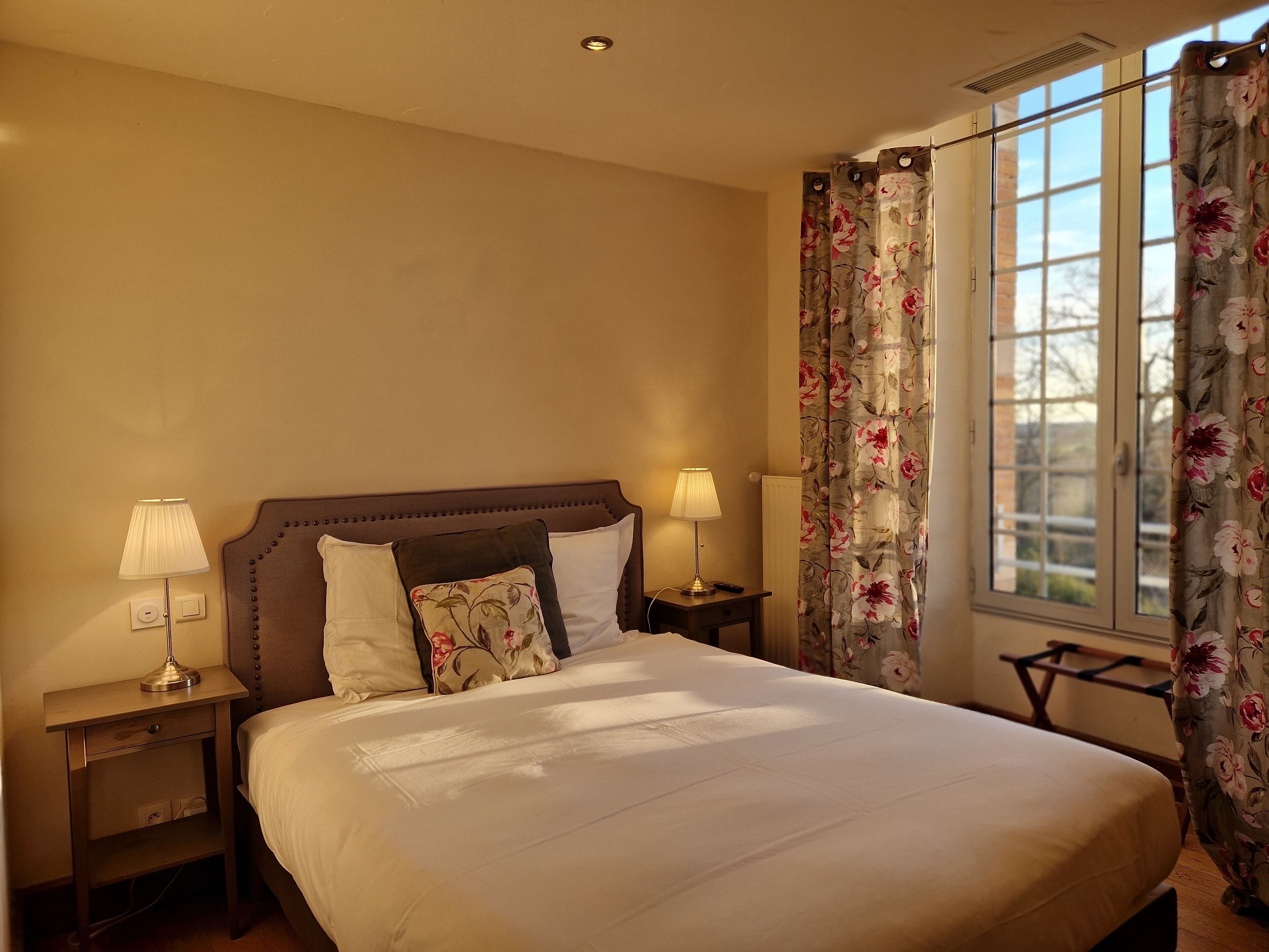 Una cama bien hecha con sábanas blancas, dos lámparas de noche, una cortina con estampado floral y vistas al exterior a través de una ventana.