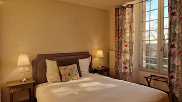 Una cama bien hecha con sábanas blancas, dos lámparas de noche, una cortina con estampado floral y vistas al exterior a través de una ventana.