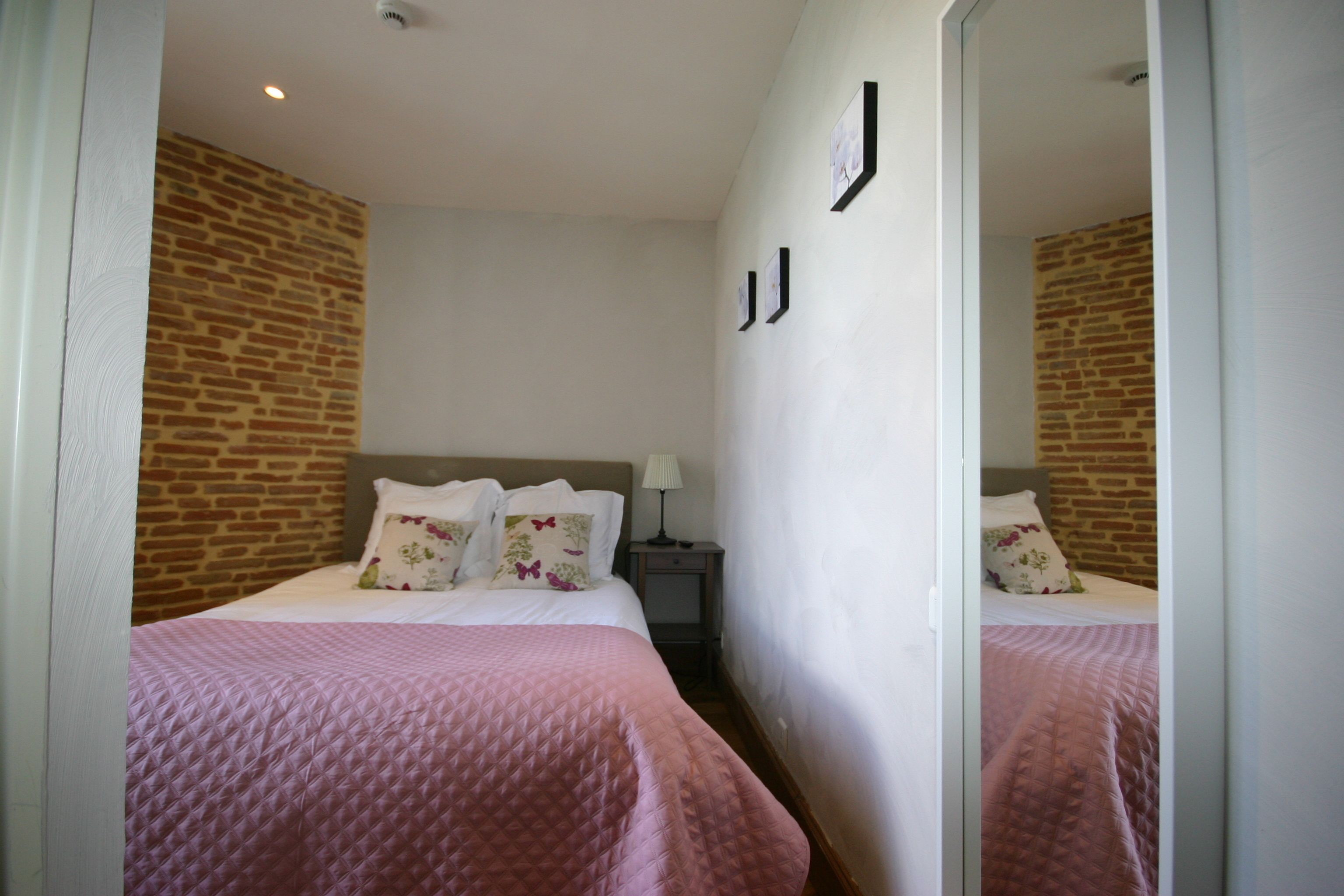 A hotel room with a brick wall, a bed with a pink quilt, a nightstand with a lamp, and a mirror on the wall.