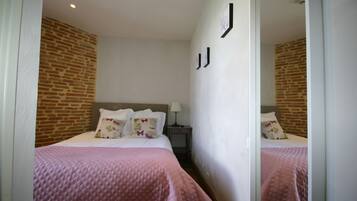A hotel room with a brick wall, a bed with a pink quilt, a nightstand with a lamp, and a mirror on the wall.