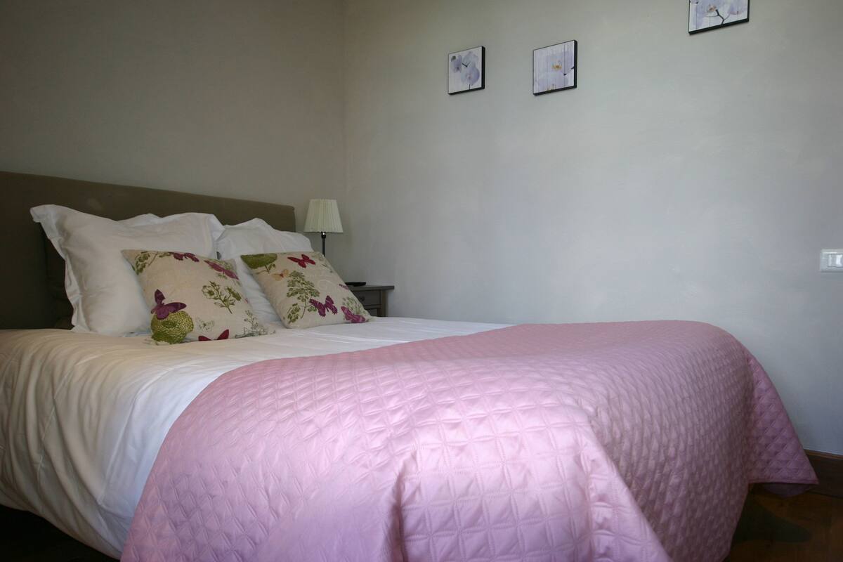 Un lit bien fait, avec une couverture rose, des oreillers blancs et un coussin à motifs floraux. Une table de chevet avec une lampe se trouve à côté du lit.