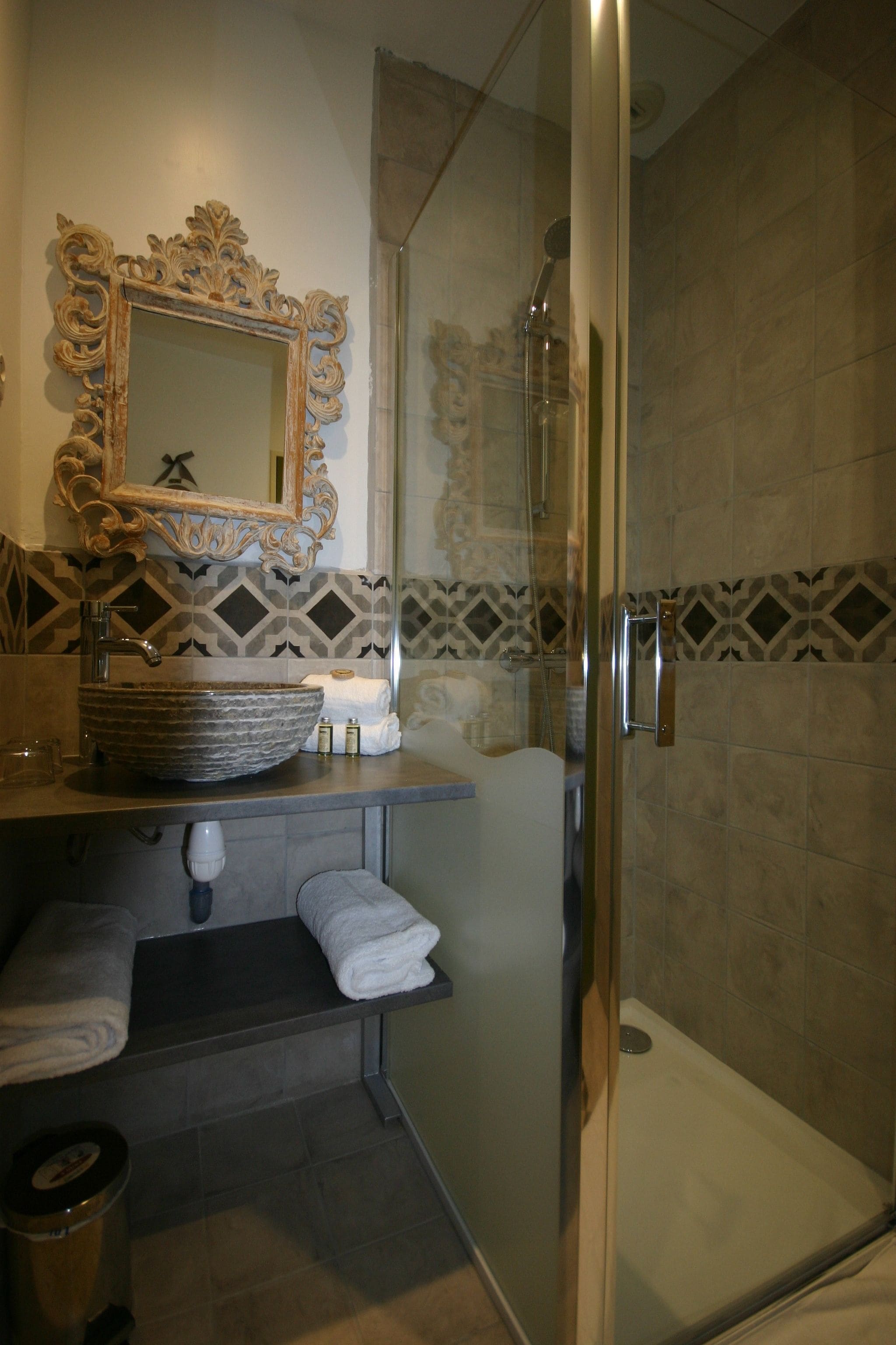 A bathroom with a glass shower enclosure, a sink with a decorative bowl, a vanity with a mirror, and patterned tiles on the walls.
