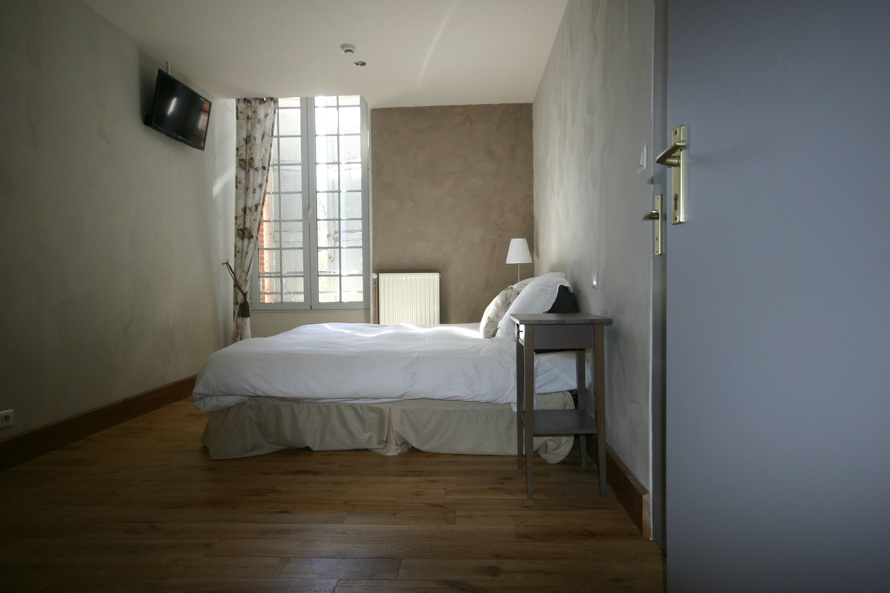 A bedroom with a bed, a nightstand, a lamp, a TV, and a window with curtains.