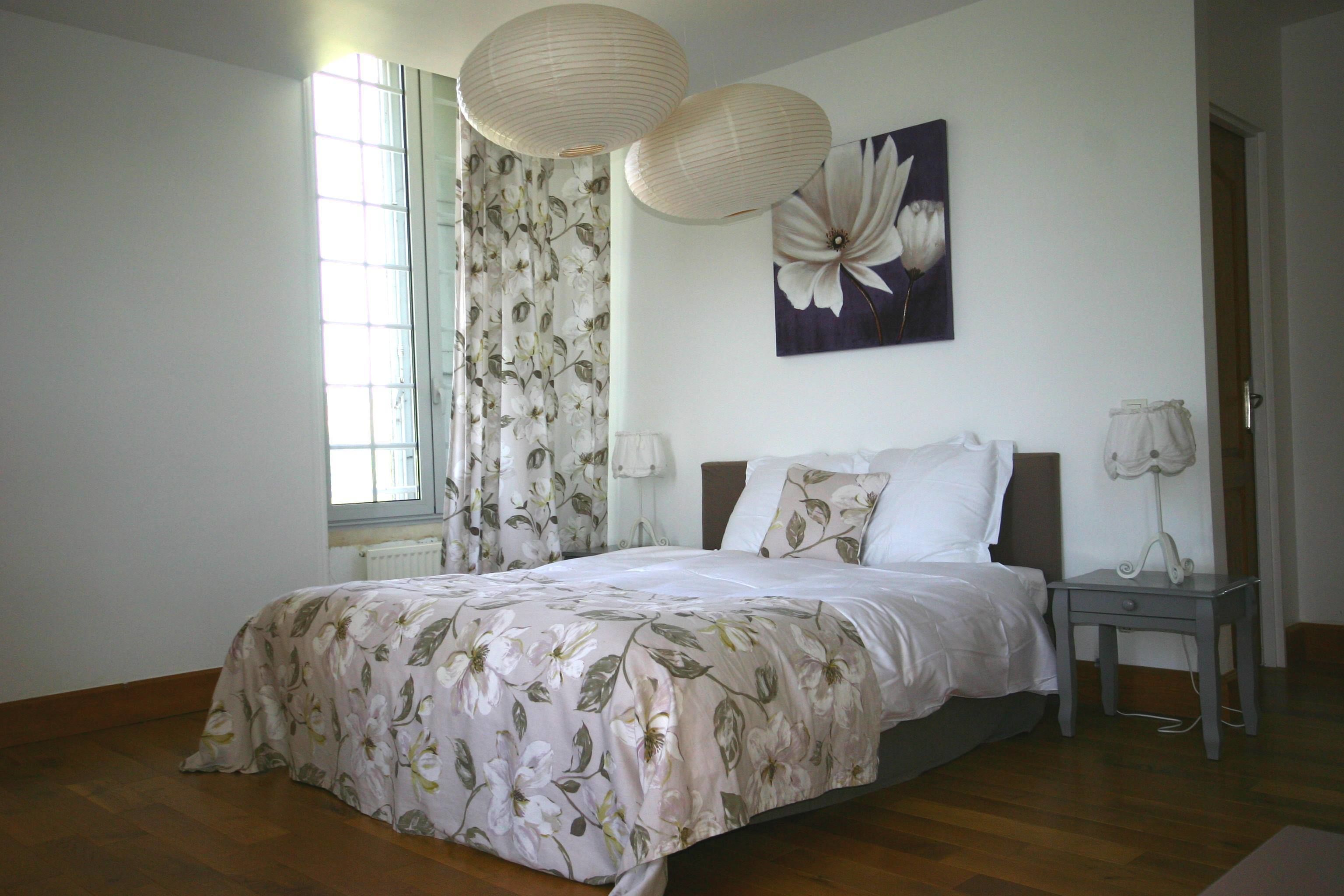 A bedroom with a floral bedspread, a large window with curtains, a nightstand with a lamp, and a wall-mounted artwork.