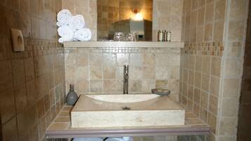 Une salle de bain avec un mur recouvert de carreaux beiges, un lavabo, un miroir et une étagère où sont posés des serviettes et des articles de toilette.