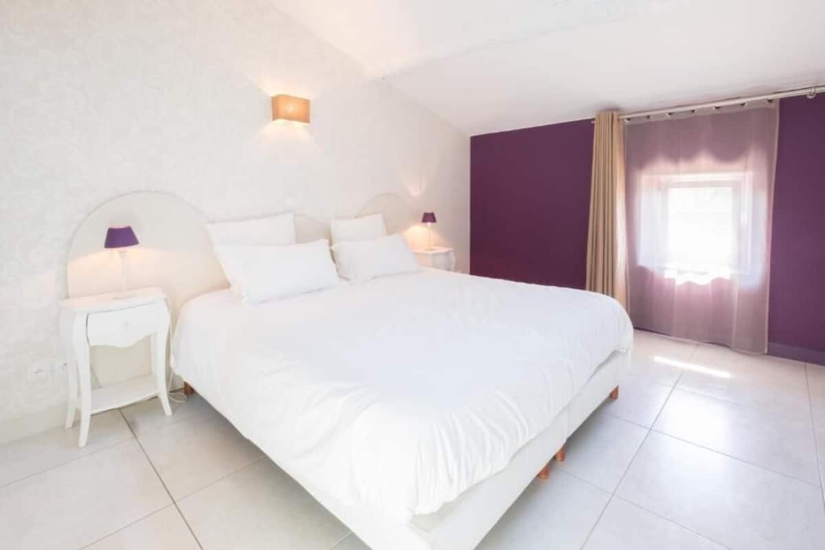 A large bed with white bedding, two bedside tables with lamps, a window with sheer curtains, and a purple accent wall.