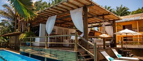 Un espace aménagé au bord de la piscine, avec une terrasse en bois, des chaises longues et un coin salon couvert équipé de parasols.