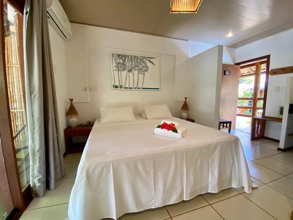 A hotel room with a large bed, a painting of palm trees, a bedside table with a lamp, and a view of the outside.