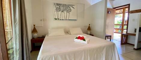 A hotel room with a large bed, a painting of palm trees, a bedside table with a lamp, and a view of the outside.