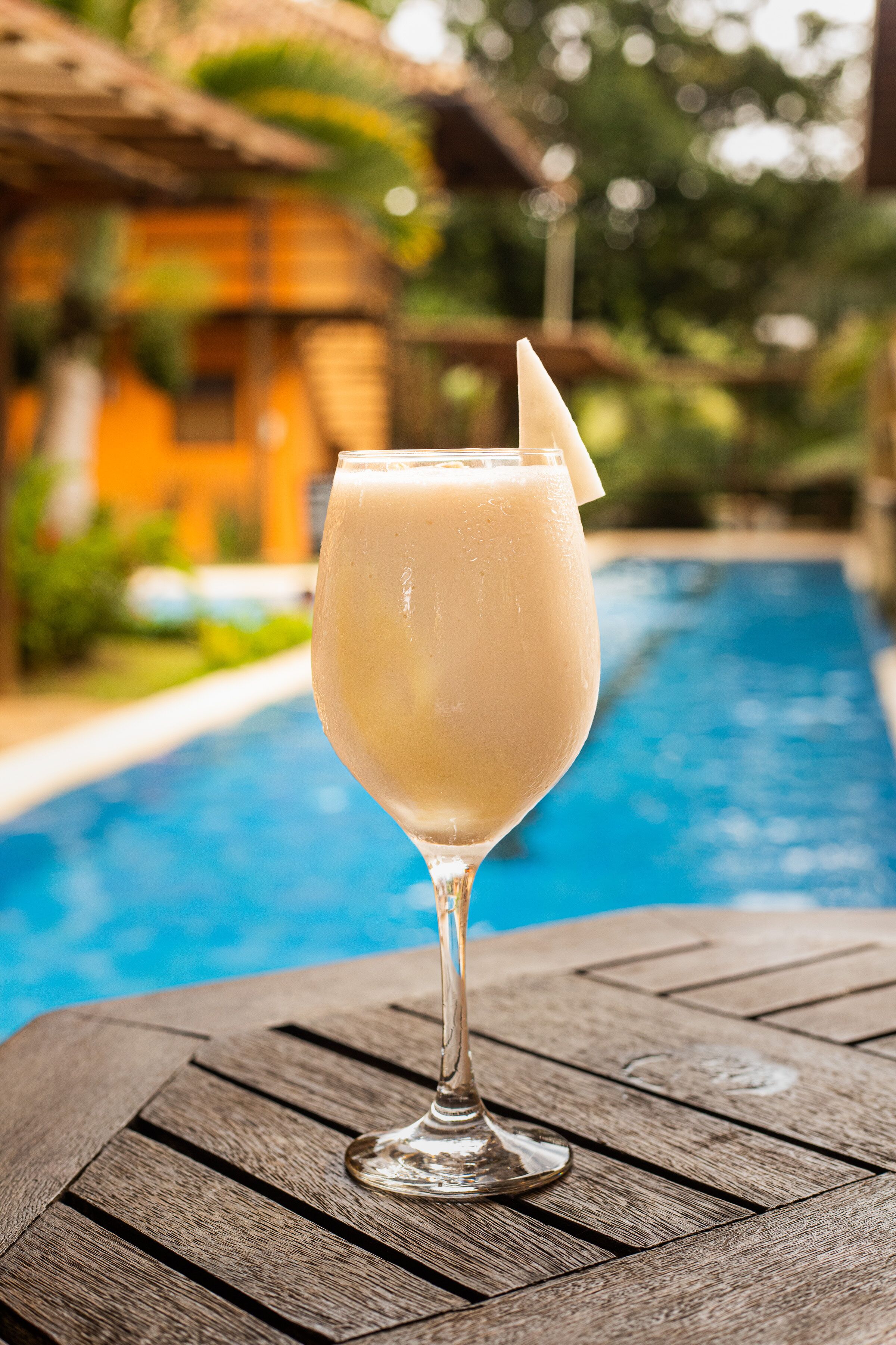 A glass of a light-colored beverage on a wooden table by a pool.