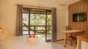 A hotel room with a bed, a table, a chair, a TV, and a view of greenery through the window.