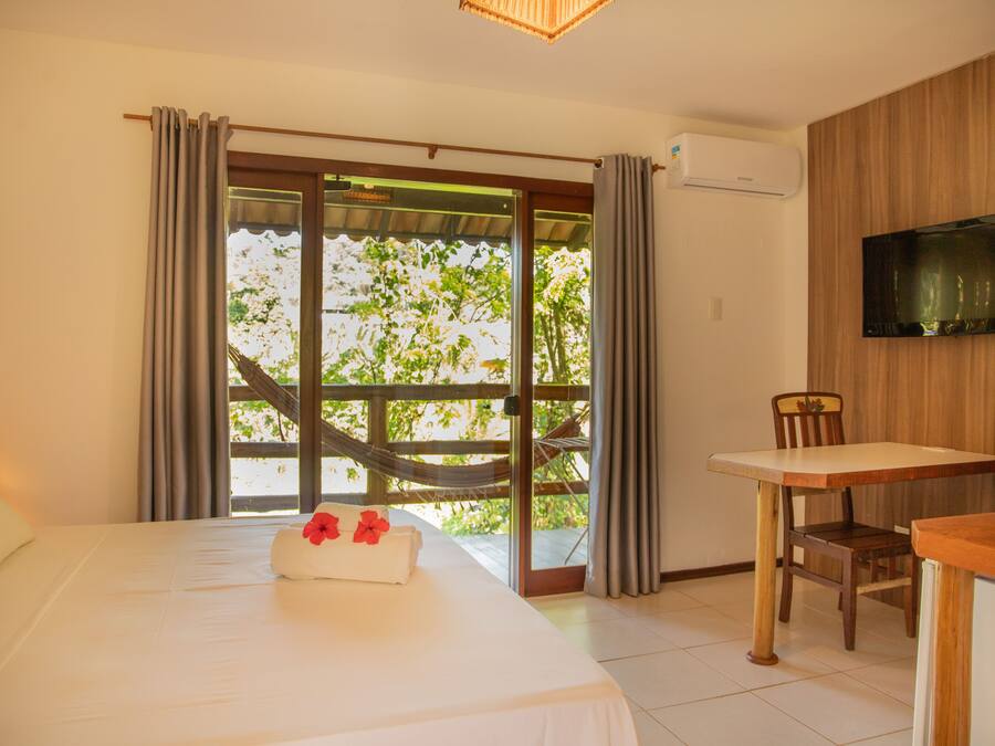 A hotel room with a bed, a table, a chair, a TV, and a view of greenery through the window.