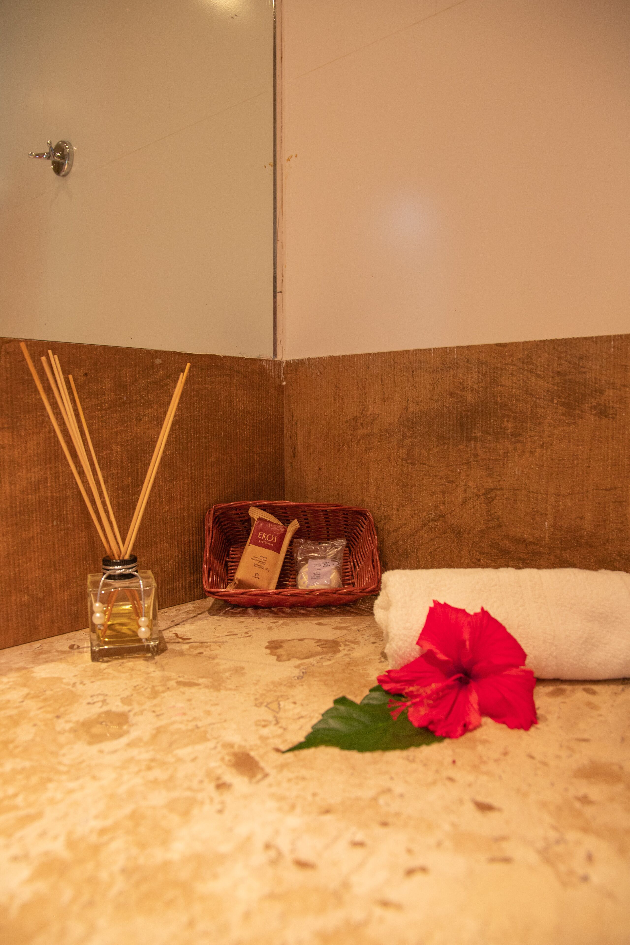 A bathroom with a marble floor, a red hibiscus flower, a glass bottle with reed sticks, and a wicker basket with toiletries.
